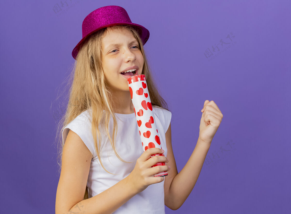 女孩美丽的小女孩戴着节日帽子拿着派对饼干用麦克风唱歌 生日派对的概念站在紫色的背景上歌手节日站