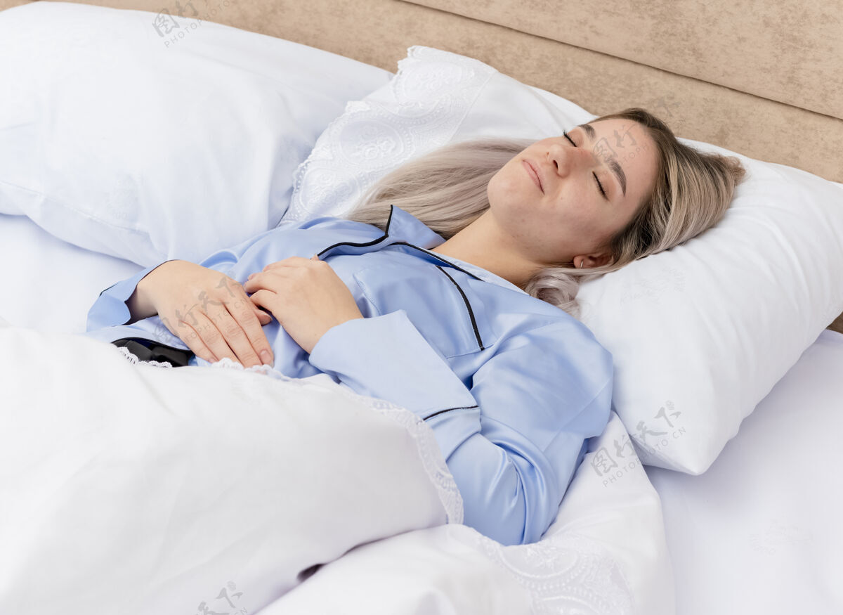 躺着穿着蓝色睡衣的年轻漂亮女人躺在床上 躺在柔软的枕头上 安详地睡在家里卧室的室内灯光背景下柔软年轻床