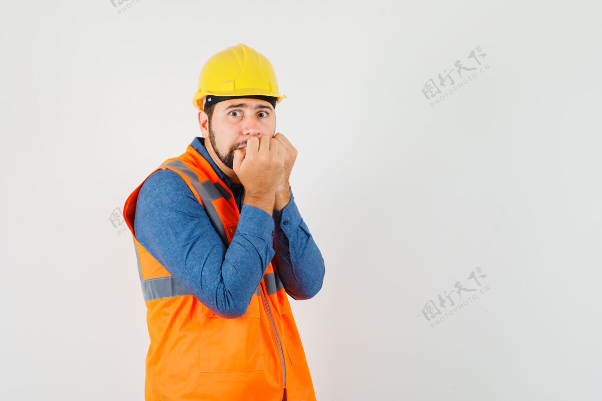 工匠年轻的建筑工人穿着衬衫 背心 头盔咬拳头的情绪和看起来害怕 前视图工作制服工具