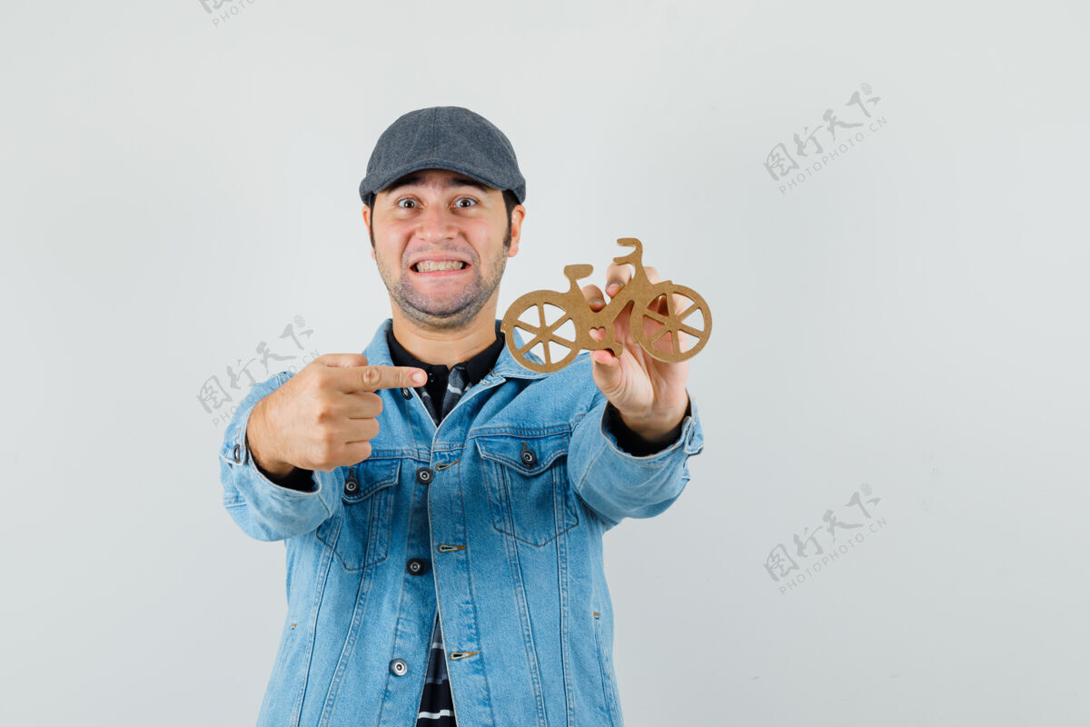 夹克一个戴帽子 穿t恤衫 穿夹克衫的年轻人指着木制玩具自行车 看上去很高兴休闲木头成功