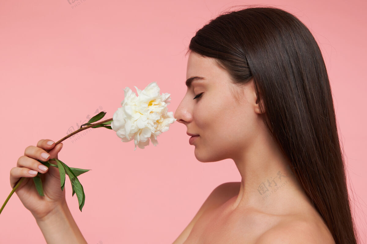 女性可爱的女人 闭着眼睛 深褐色的长发 健康的皮肤 用花触摸鼻子 闻着香味特写站在粉红色的墙壁上成人健康20多岁
