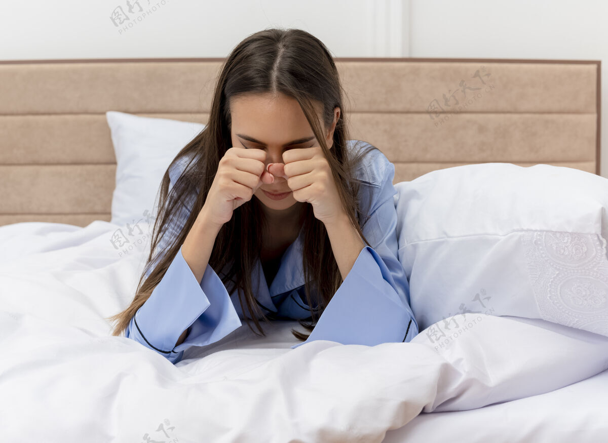 睡衣穿着蓝色睡衣的年轻美女躺在床上揉眼睛打呵欠感觉晨曦疲惫在卧室的室内灯光背景下感觉眼睛打哈欠