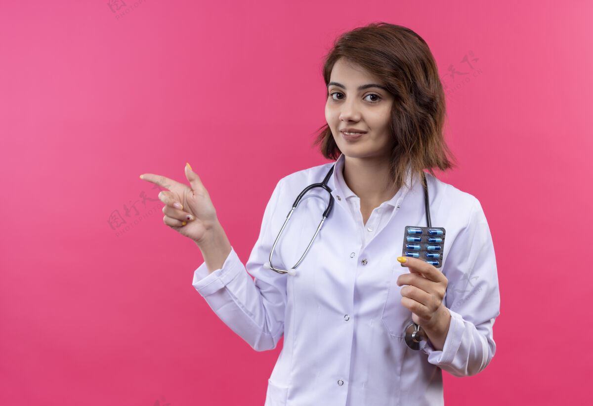 年轻身穿白大褂的年轻女医生拿着听诊器拿着水泡 拿着药丸 手指指向一旁微笑着手指水泡女人