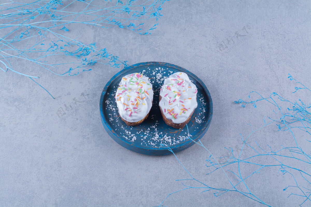 餐盘两个新鲜的甜纸杯蛋糕 蓝色木板上有五颜六色的洒点和奶油美味洒水烘焙