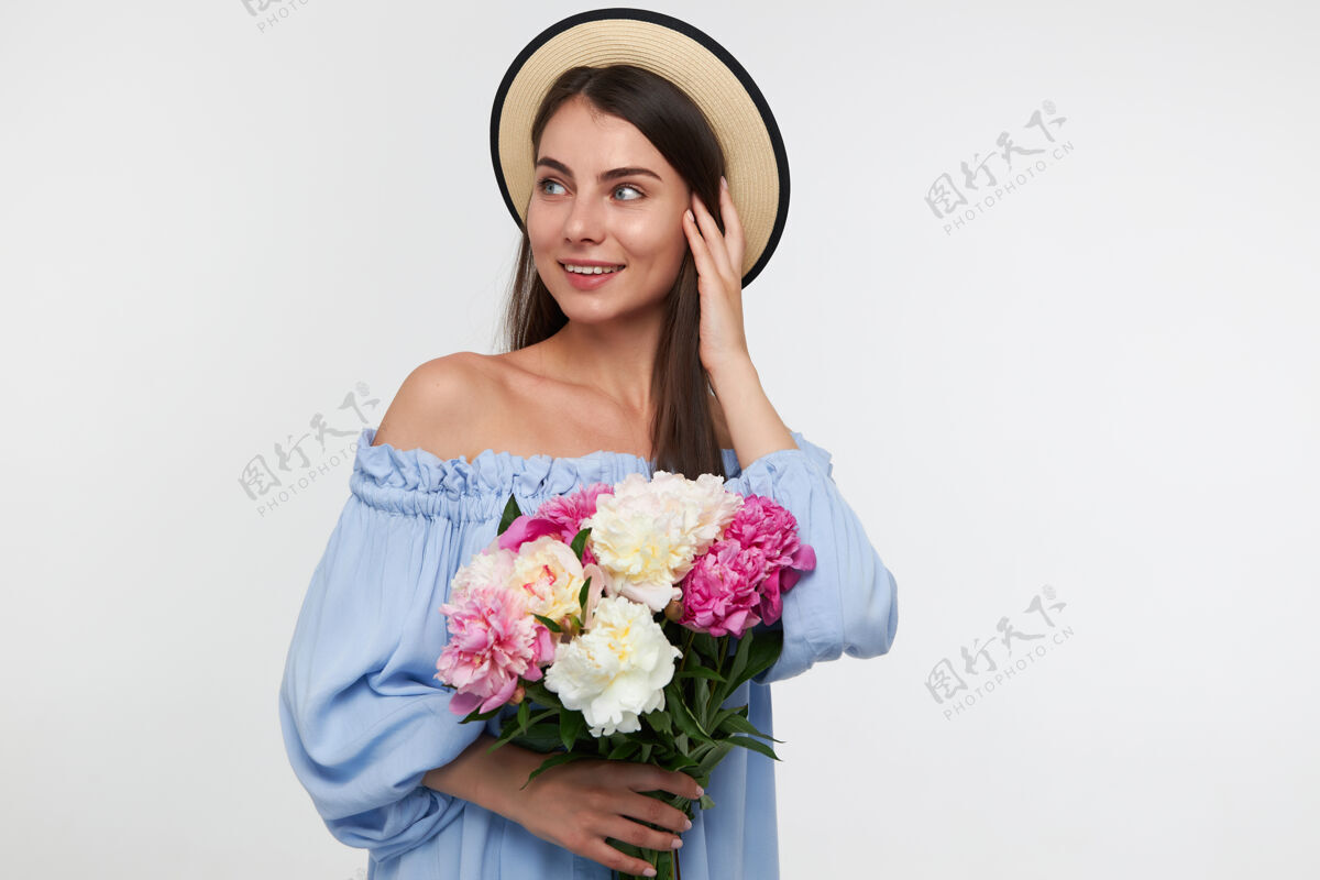 青少年十几岁的女孩 快乐的女人 深色长发戴着帽子 穿着蓝色漂亮的裙子拿着一束花 抚摸着头发在白墙上的复制空间看着左边皮肤装花