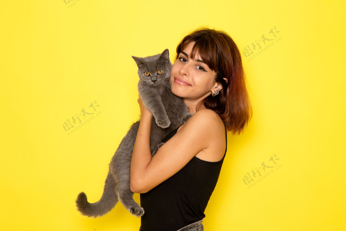 年轻身穿黑色衬衫的年轻女性在黄色墙壁上微笑着抱着小猫的正面视图衬衫抱着前面