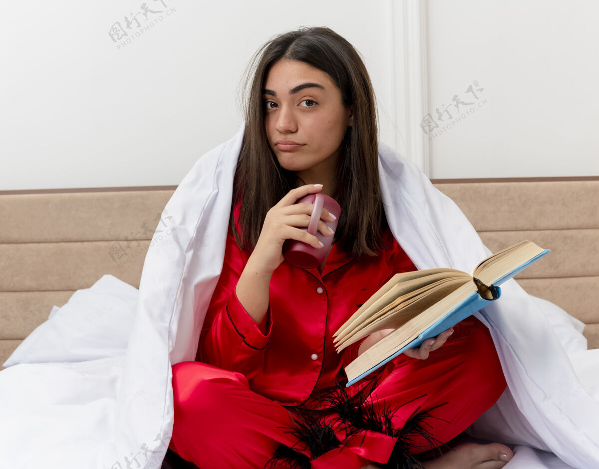 怀疑穿着红色睡衣的年轻美女坐在床上 裹着毯子 端着一杯咖啡和一本书 看着摄像机 脸上带着怀疑的表情 在卧室内部的灯光背景下相机脸咖啡