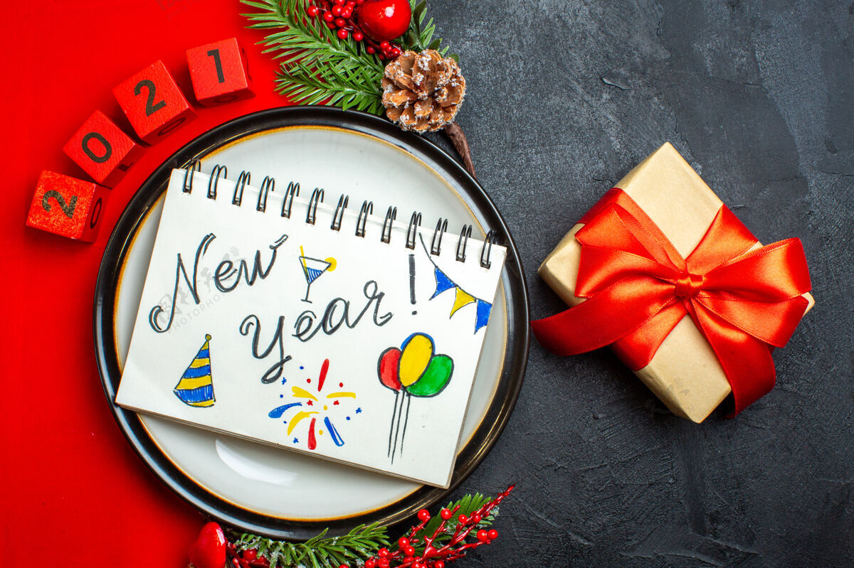 图纸新年背景俯视图 笔记本上有新年图纸 餐盘上有装饰配件 红餐巾上有杉木树枝和数字 黑桌子上有礼物餐盘顶部树枝
