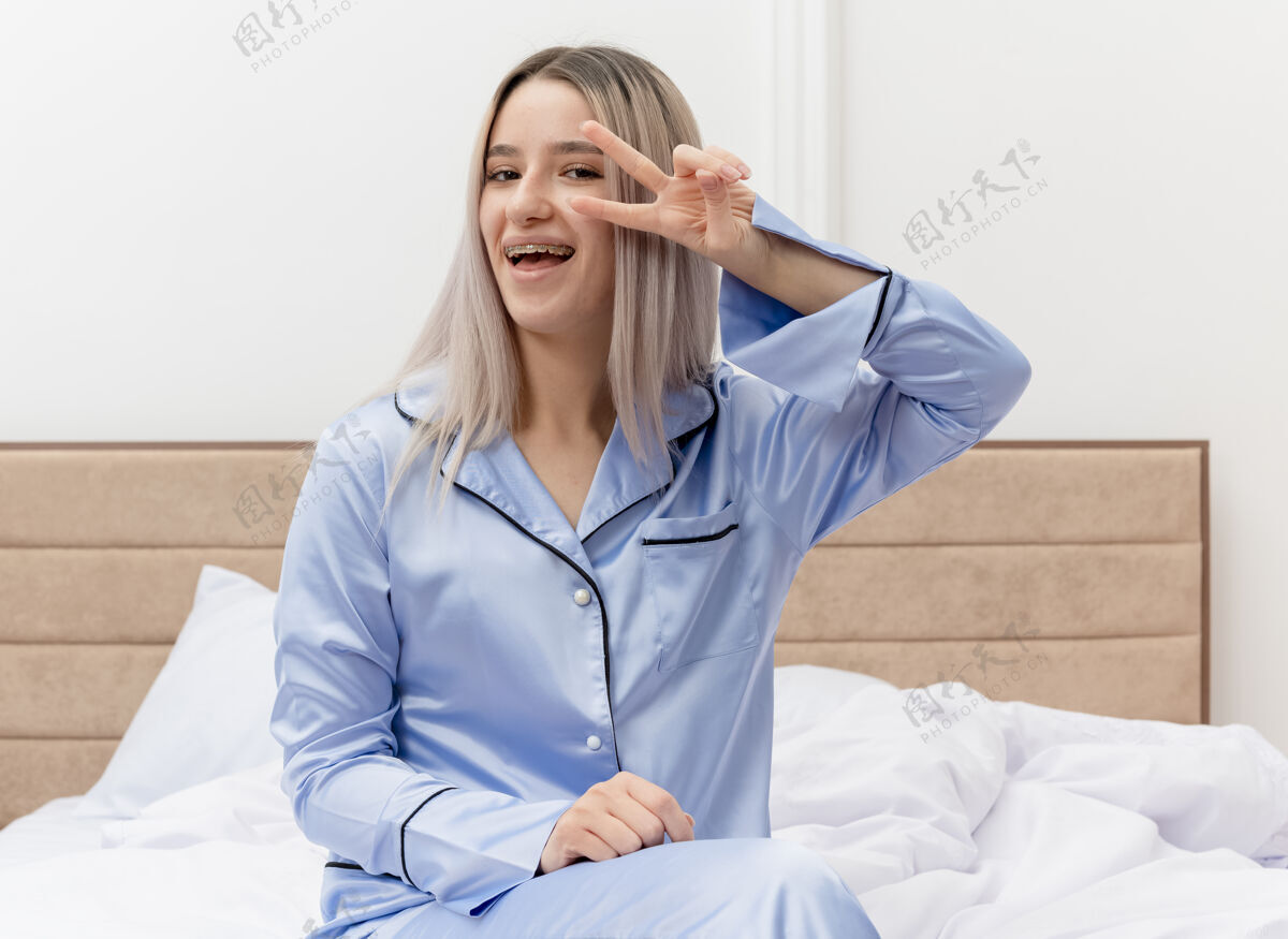 Vsign穿着蓝色睡衣的年轻漂亮女人坐在床上 微笑着 在卧室的室内灯光背景下 带着微笑 露出v字标志微笑室内卧室