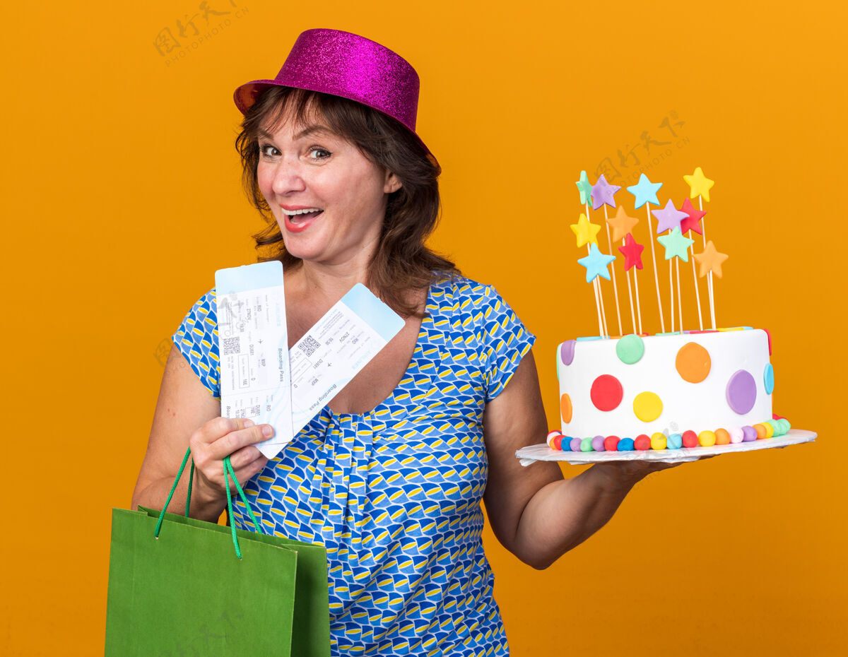 机票快乐快乐的中年妇女戴着聚会帽 手里拿着带礼物的纸袋 手里拿着生日蛋糕和机票抱票生日