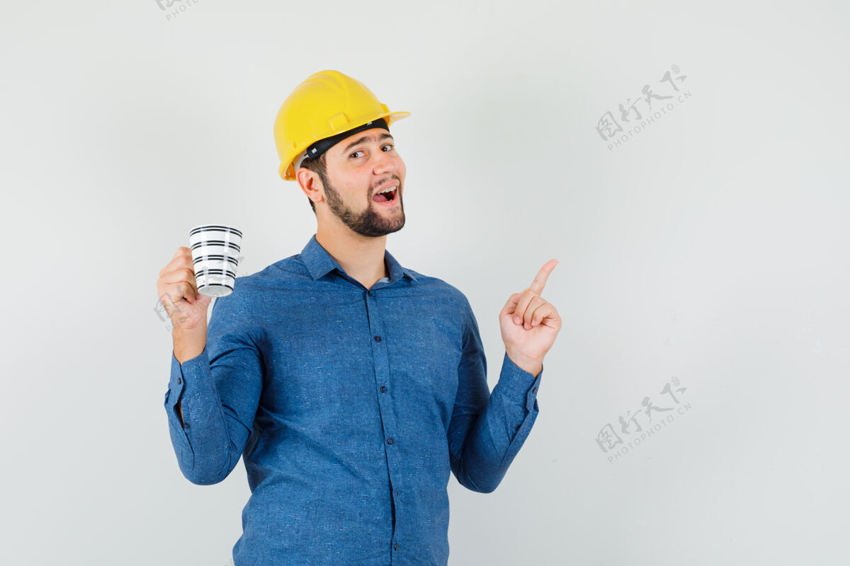 休闲年轻的男性指着头 拿着一杯咖啡 穿着衬衫 戴着头盔 看上去很高兴正面图成人站立帅气