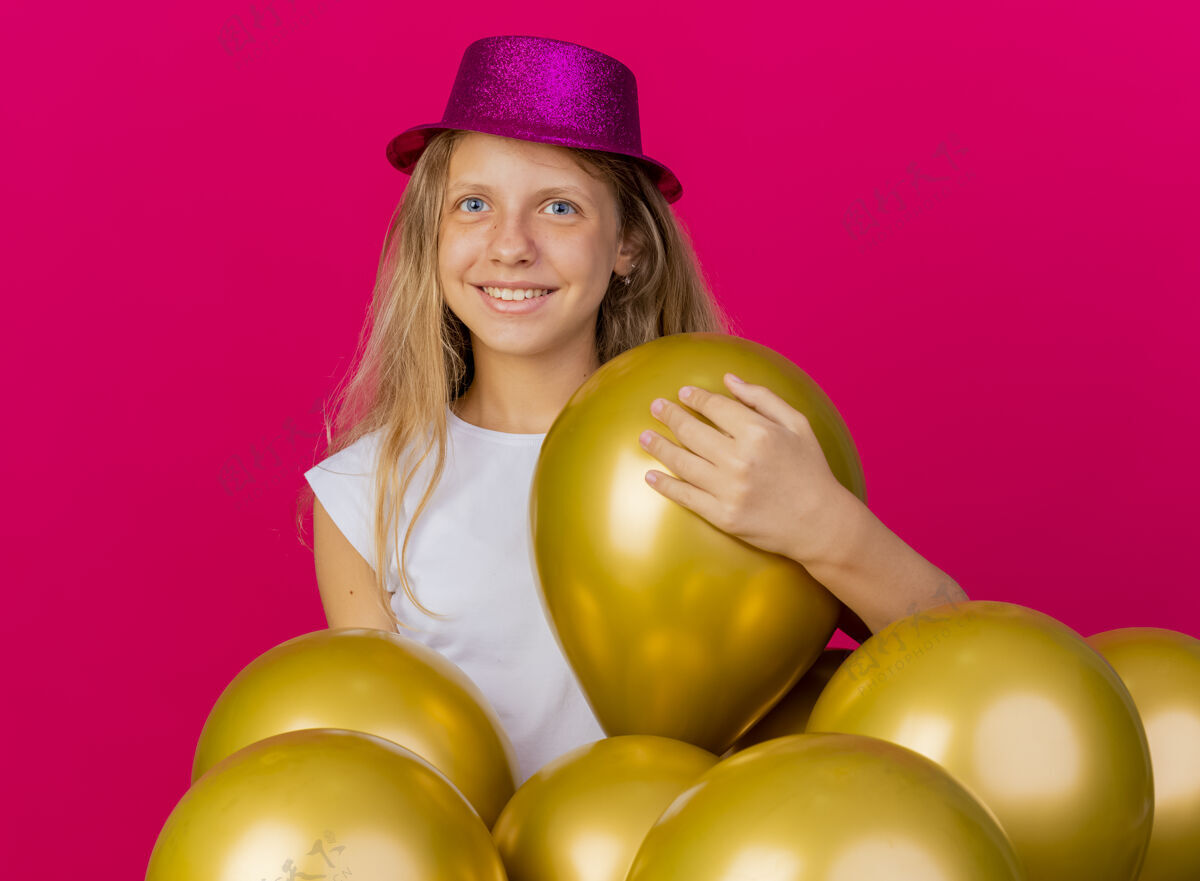 脸戴着节日礼帽的快乐可爱的小女孩和一群巴隆人看着镜头笑脸盈盈 粉色背景下站着生日派对的概念节日欢呼微笑