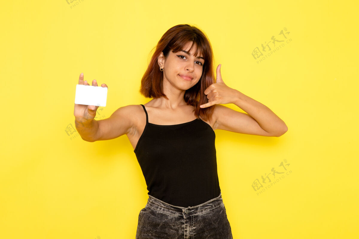 漂亮身穿黑色衬衫和灰色牛仔裤的年轻女性在黄色墙壁上手持白色卡片的正面视图姿势女孩衬衫