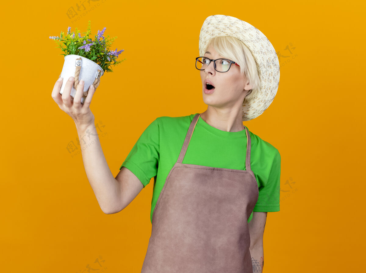 帽子围裙上留着短发 戴着帽子 手里拿着盆栽植物的年轻园丁站在橙色的背景下 惊讶地看着它期待植物举行