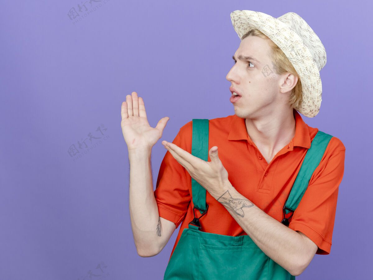 帽子年轻的园丁 穿着连体衣 戴着帽子 手放在一边 站在紫色的背景上 困惑地看着看花园男人