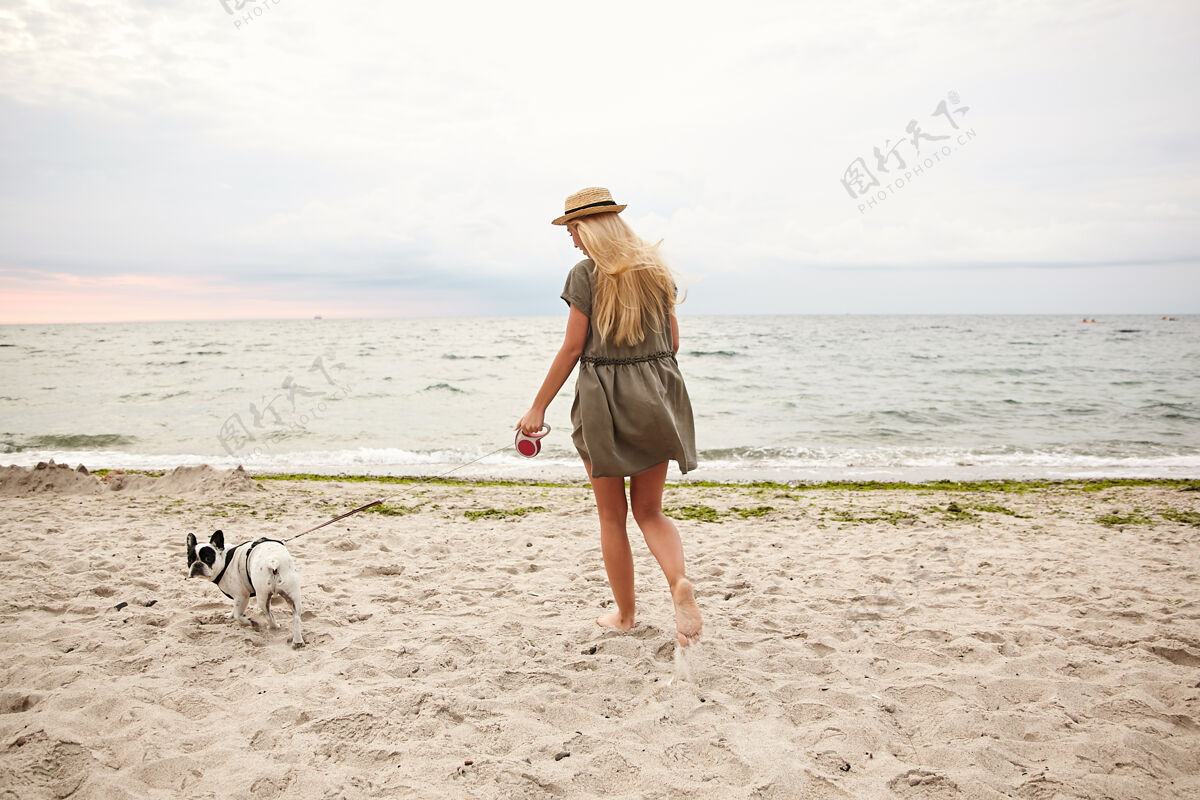 情绪户外照片 苗条的年轻女性 金色长发 穿着夏装 头戴水手帽 在灰蒙蒙的阴天 牵着狗在沙滩上散步白色阴天姿势