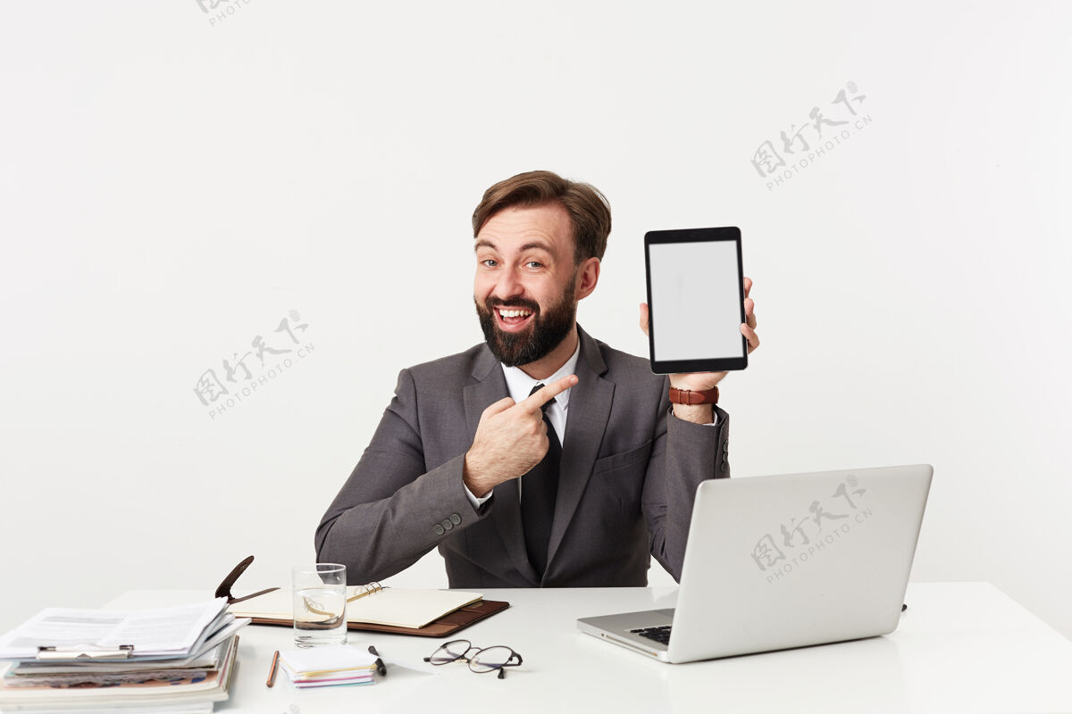 便条喜气洋洋的年轻人 留着胡子 深色头发 时髦的发型 用食指指着手中的平板电脑 面带微笑 身着灰色西装 坐在白墙上的桌子旁衣服男人西装