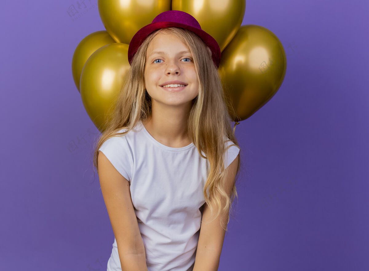 微笑戴着节日礼帽的漂亮小女孩和一群巴隆人看着镜头 微笑着快乐和积极 紫色背景下的生日派对概念站节日漂亮