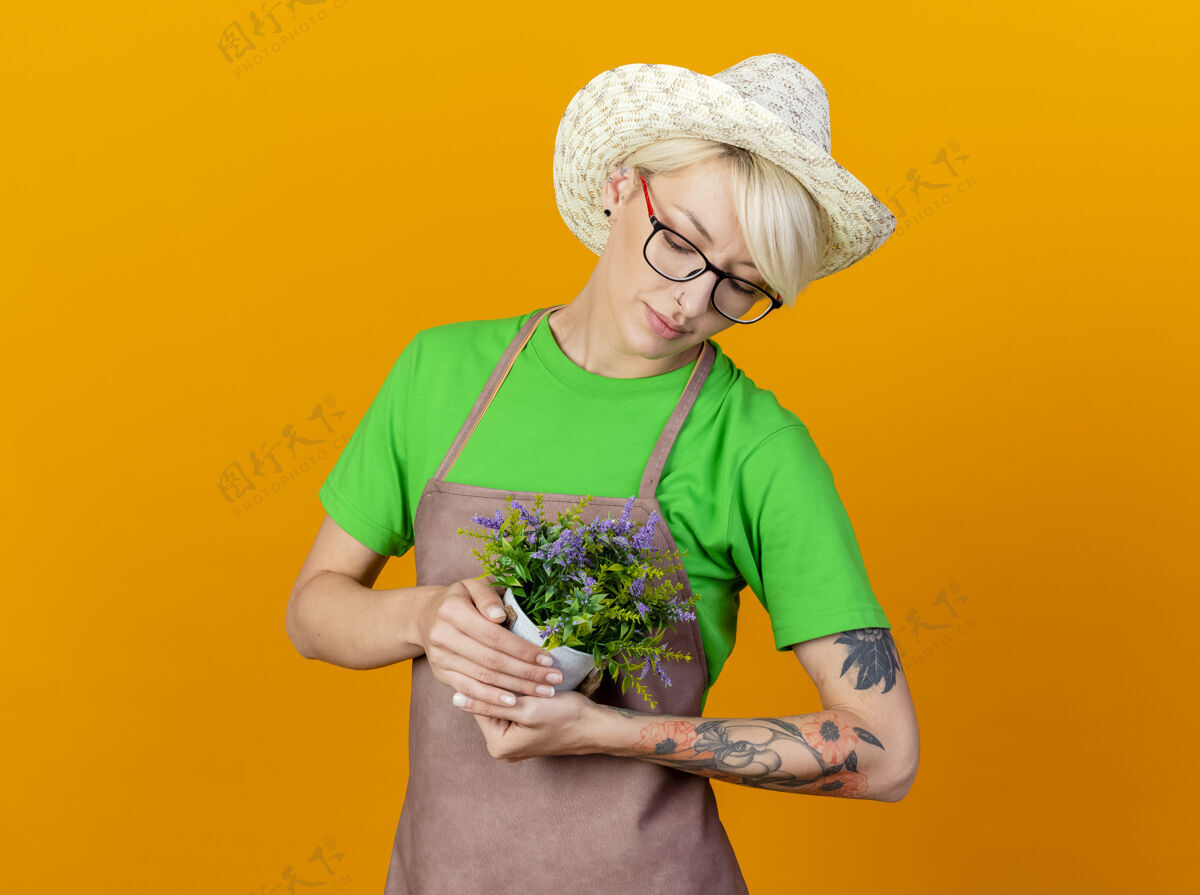 橙色一位年轻的园丁 围裙上留着短发 戴着帽子 手里拿着盆栽植物 站在橙色的背景下 带着爱看着它头发围裙植物