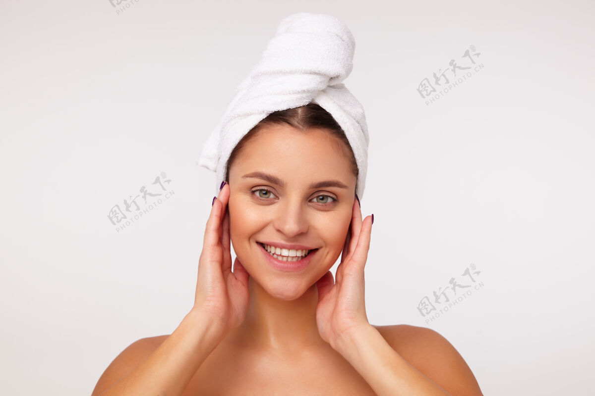 裸体一位迷人的年轻女士的特写镜头 黑色头发裹在浴巾里 举手触摸着她的脸 脸上洋溢着灿烂的笑容 站在白色的背景下微笑白色年轻