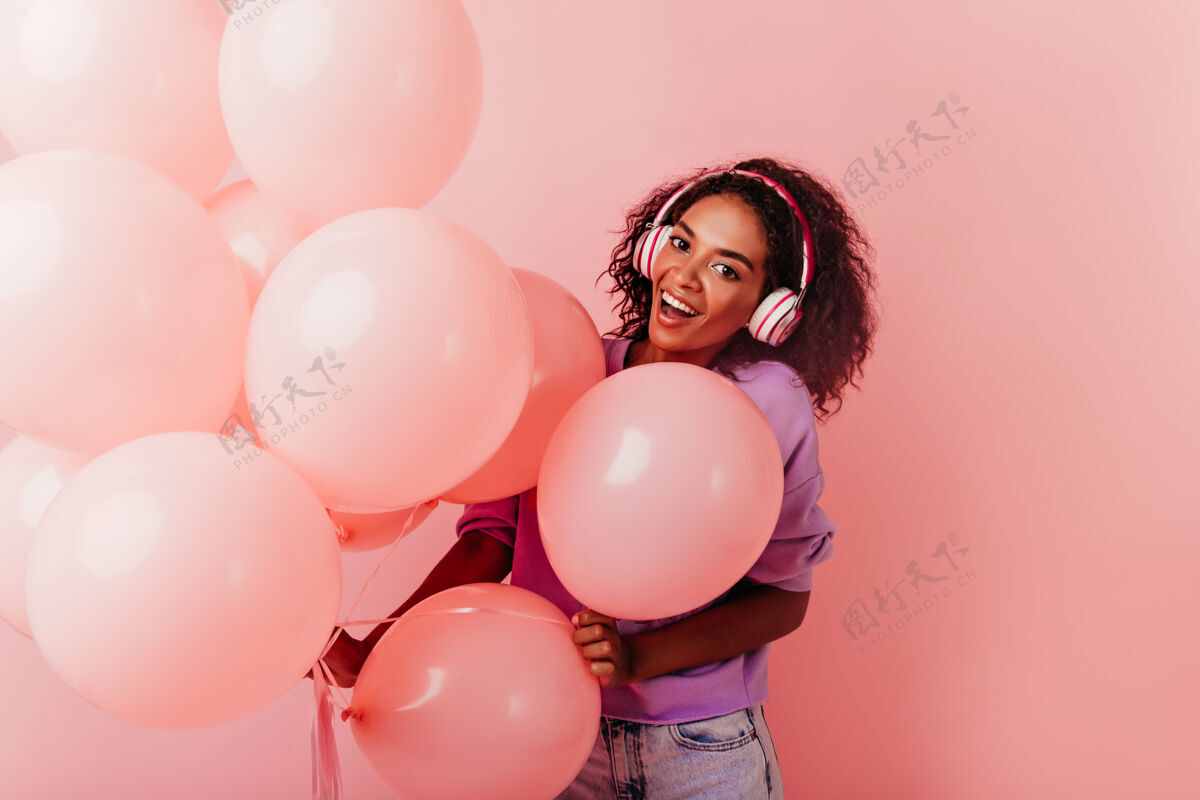 年轻人戴着大耳机的兴奋的生日女孩与气球合影在聚会上听音乐的优雅的非洲女士非洲惊喜积极