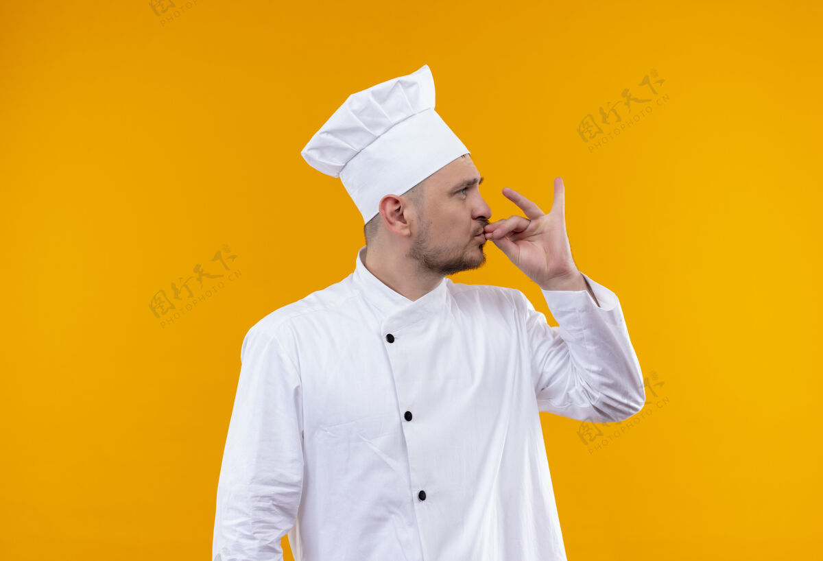 橙色年轻帅气的厨师穿着厨师制服做着美味的手势 看着旁边孤立的橙色空间手势年轻制服