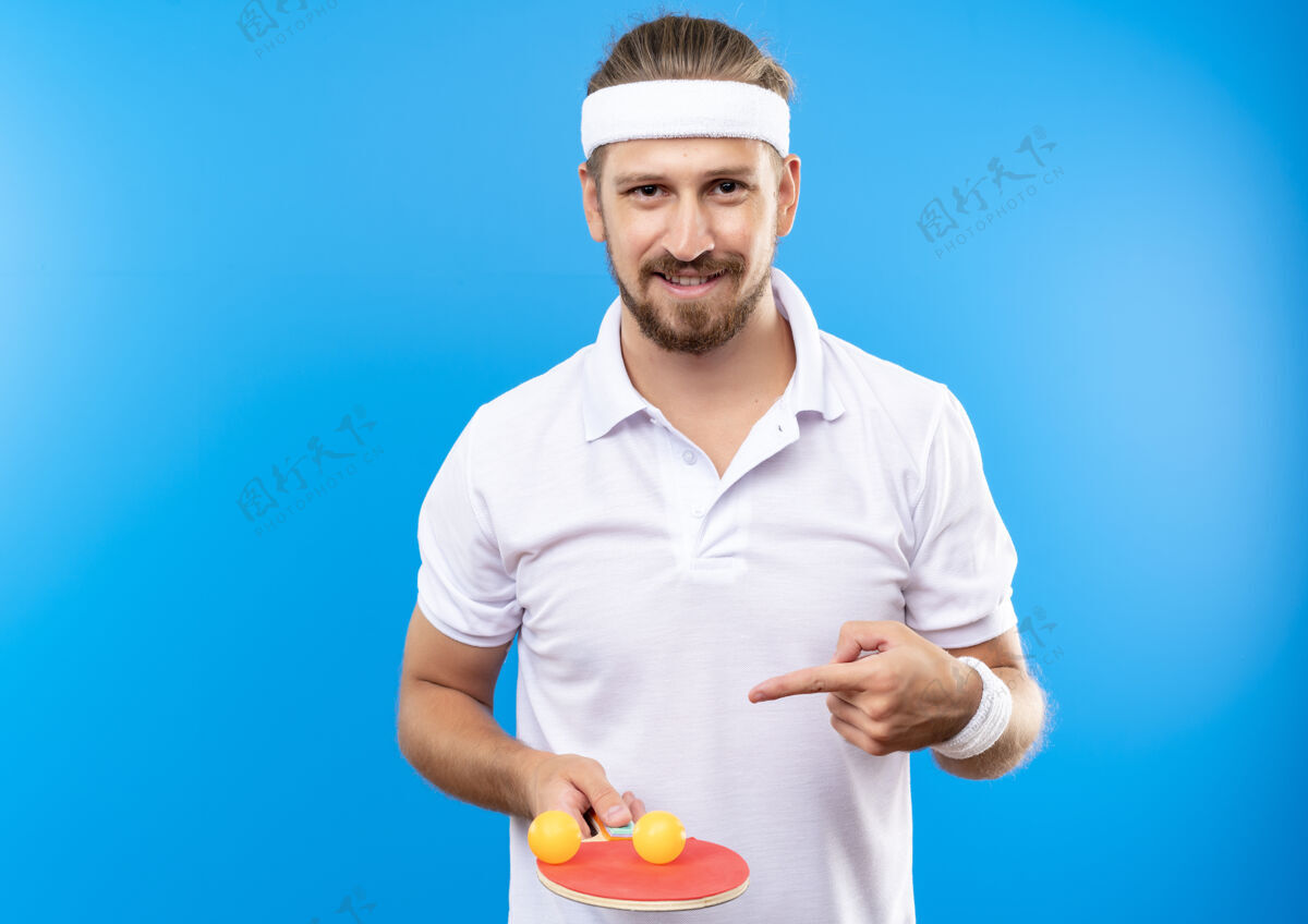 头带面带微笑的年轻英俊的运动型男子戴着头带和腕带拿着并指着乒乓球拍 球拍上的球被隔离在蓝色的空间里微笑复制腕带
