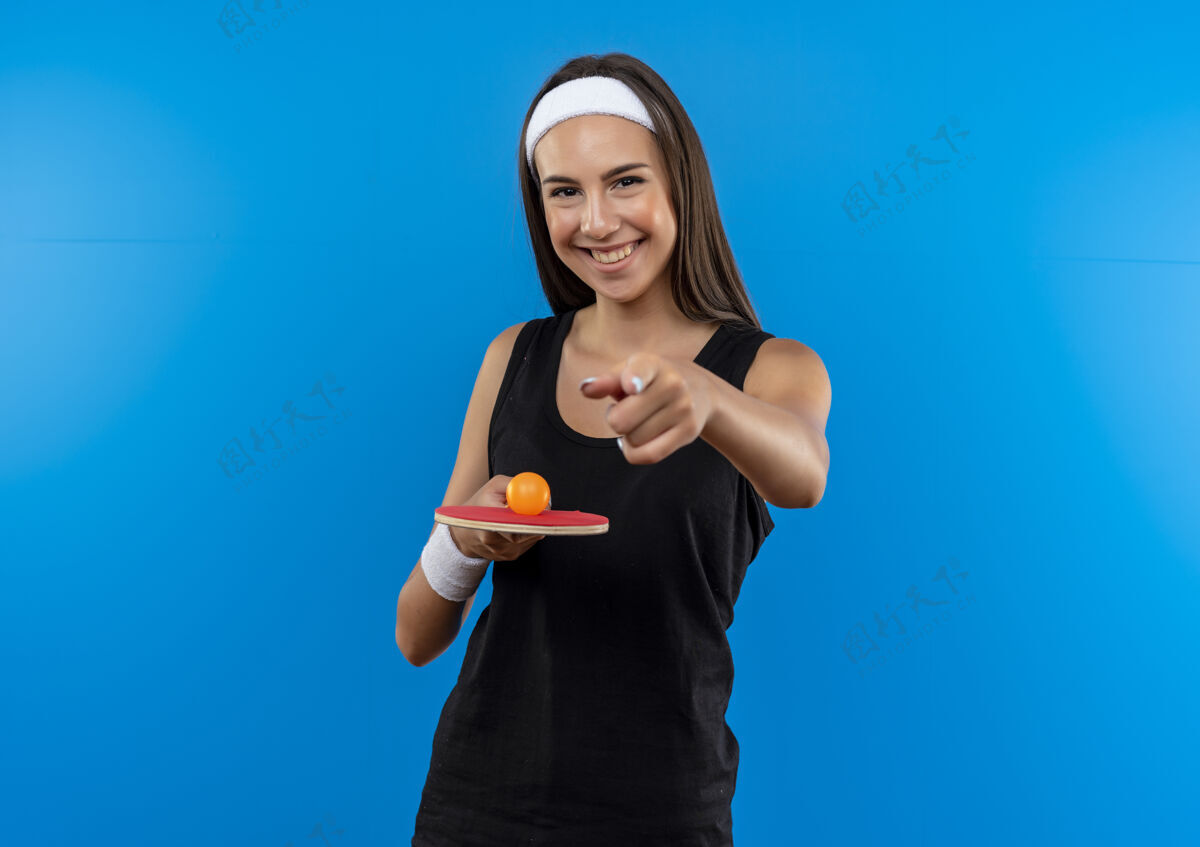 微笑微笑的年轻漂亮的运动女孩戴着头带和腕带拿着乒乓球拍 球指向蓝色空间女孩头带运动