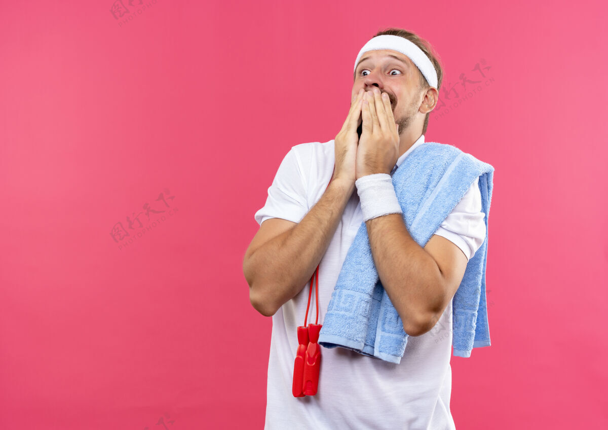 运动被吓到的年轻英俊的运动型男子戴着头带和腕带双手放在嘴上看着一边 肩上系着跳绳和毛巾隔离在粉色的空间里穿粉红色空间
