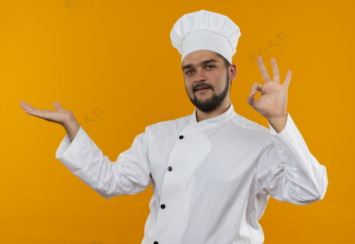 请身着厨师制服的年轻男厨师很高兴地在橙色的空间里展示了空手和“做得好”的标志制服标志手