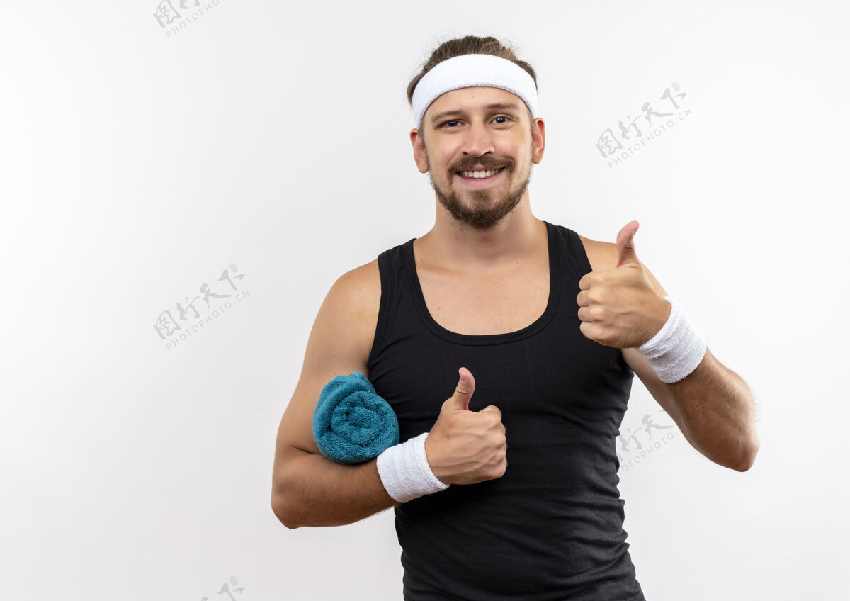 男人面带微笑的年轻帅气的运动型男人戴着头带和手环拿着毛巾竖起大拇指孤立地站在白色的空间里向上头带穿