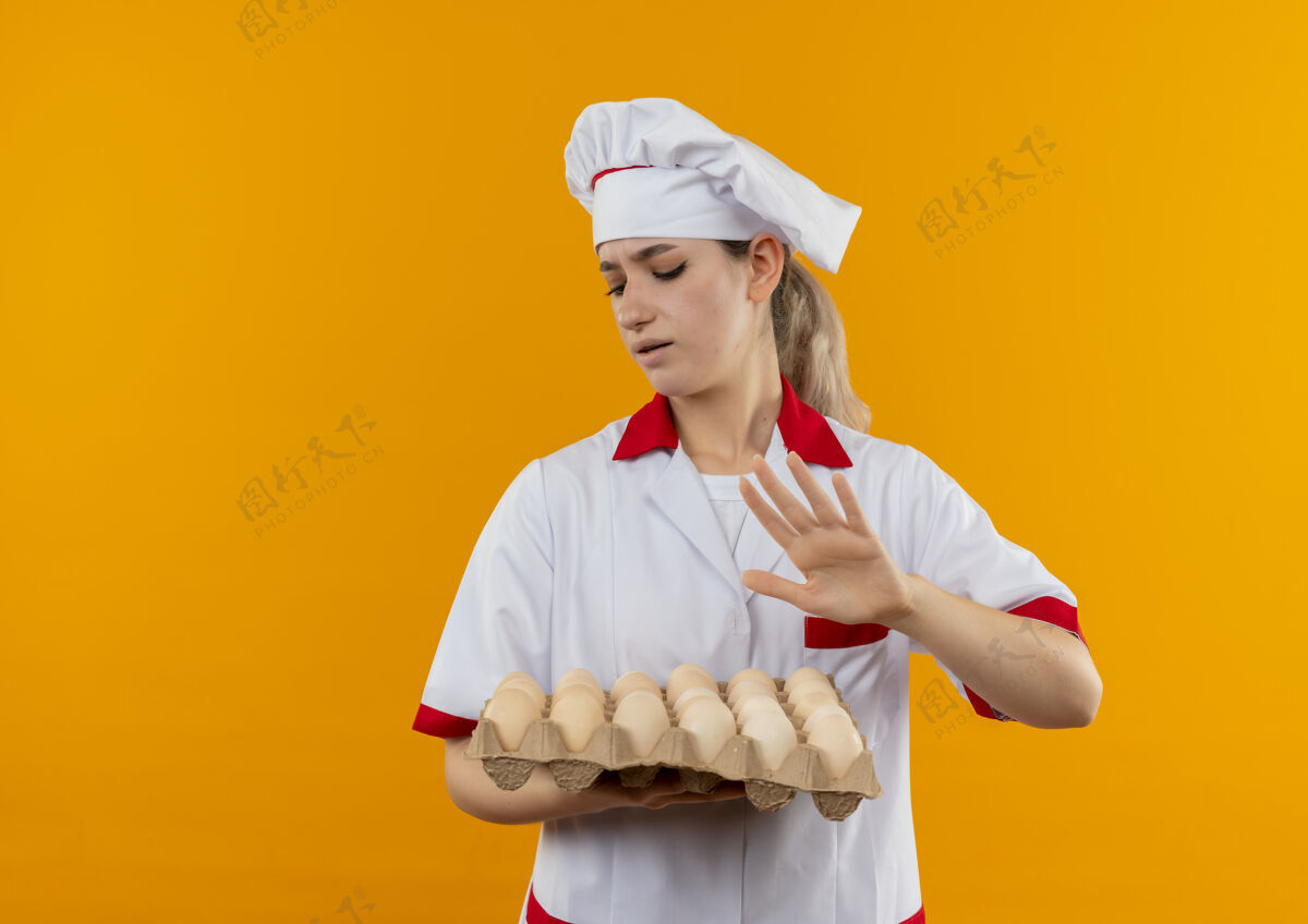 未发布年轻漂亮的厨师 穿着厨师制服 拿着一盒鸡蛋 在橙色的空间里做着不孤立的手势厨师年轻手势