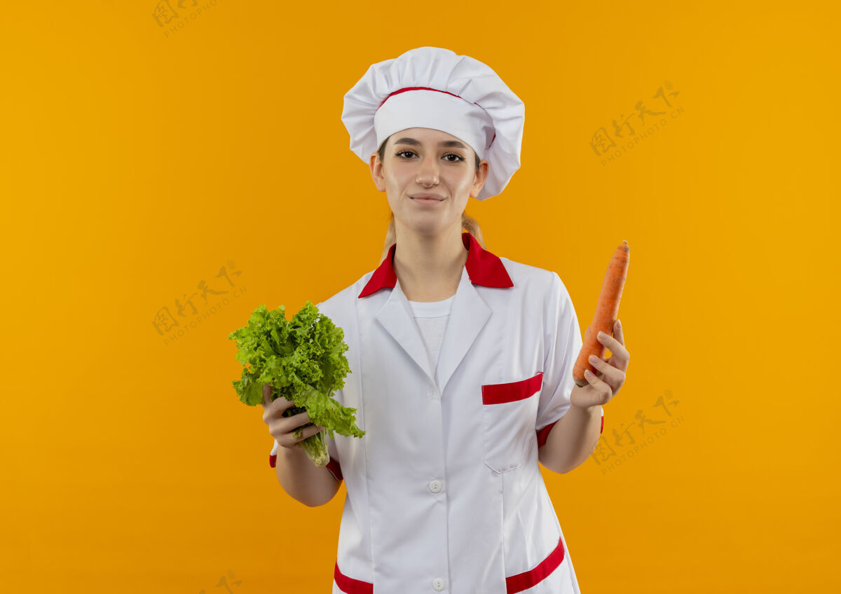 生菜年轻漂亮的厨师穿着厨师制服 手里拿着莴苣和胡萝卜 把它们隔离在橙色的空间里制服漂亮胡萝卜