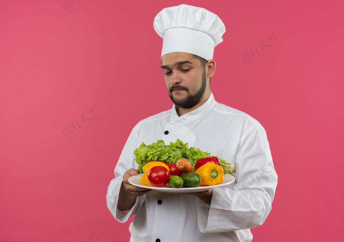 复制体贴的年轻男厨师穿着厨师制服 拿着盘子看着孤立在粉红色空间里的蔬菜体贴男性持有