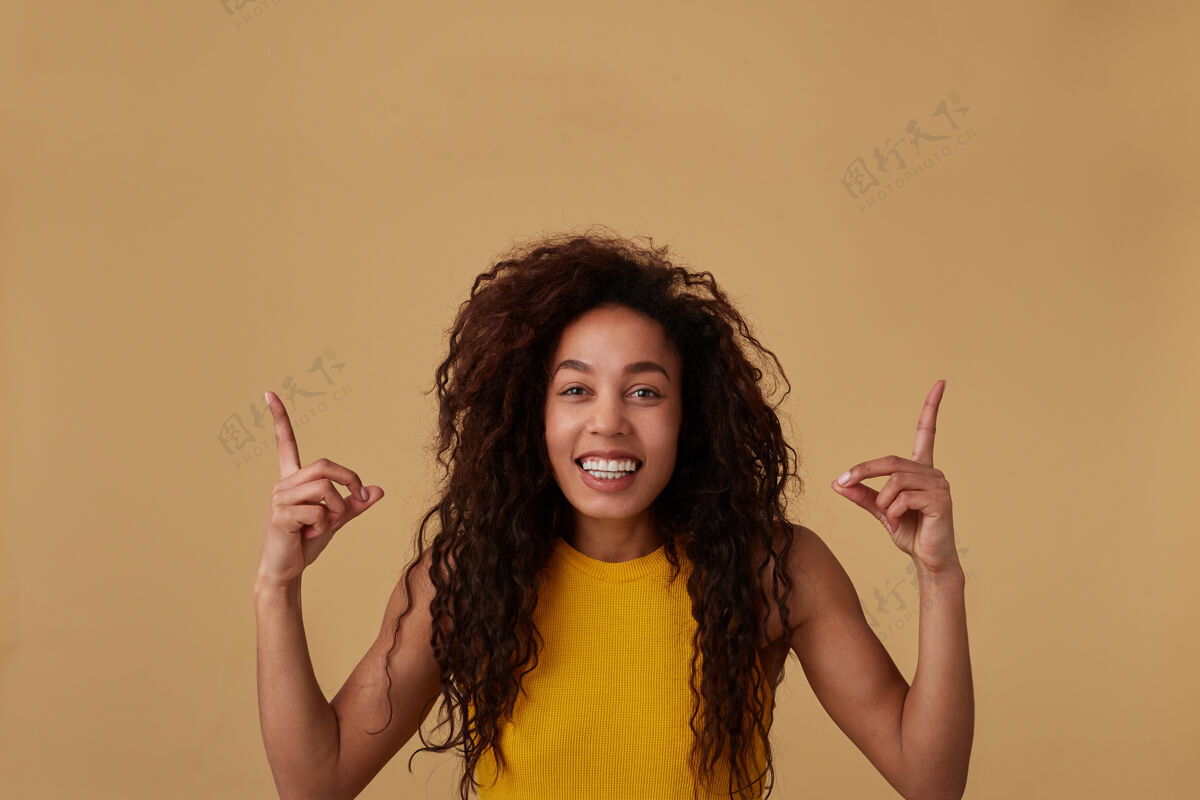 黄色快乐的年轻可爱的黑发卷曲的深色皮肤女人的肖像画自然化妆保持食指抬起 同时向上显示和微笑广泛 隔离在米色高兴女性卷发