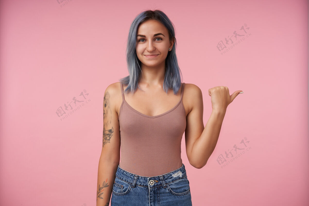 正面积极向上的年轻可爱的纹身女士 蓝色短发 微笑着 举手在一旁 穿着休闲装的粉红色被孤立发型室内米色