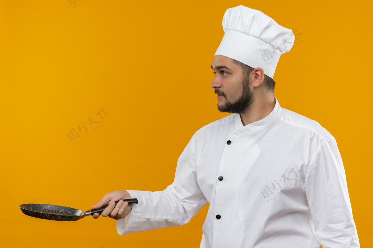 厨师身穿厨师制服的年轻男厨师手拿煎锅 看着橙色空间上孤立的一面看拿着男