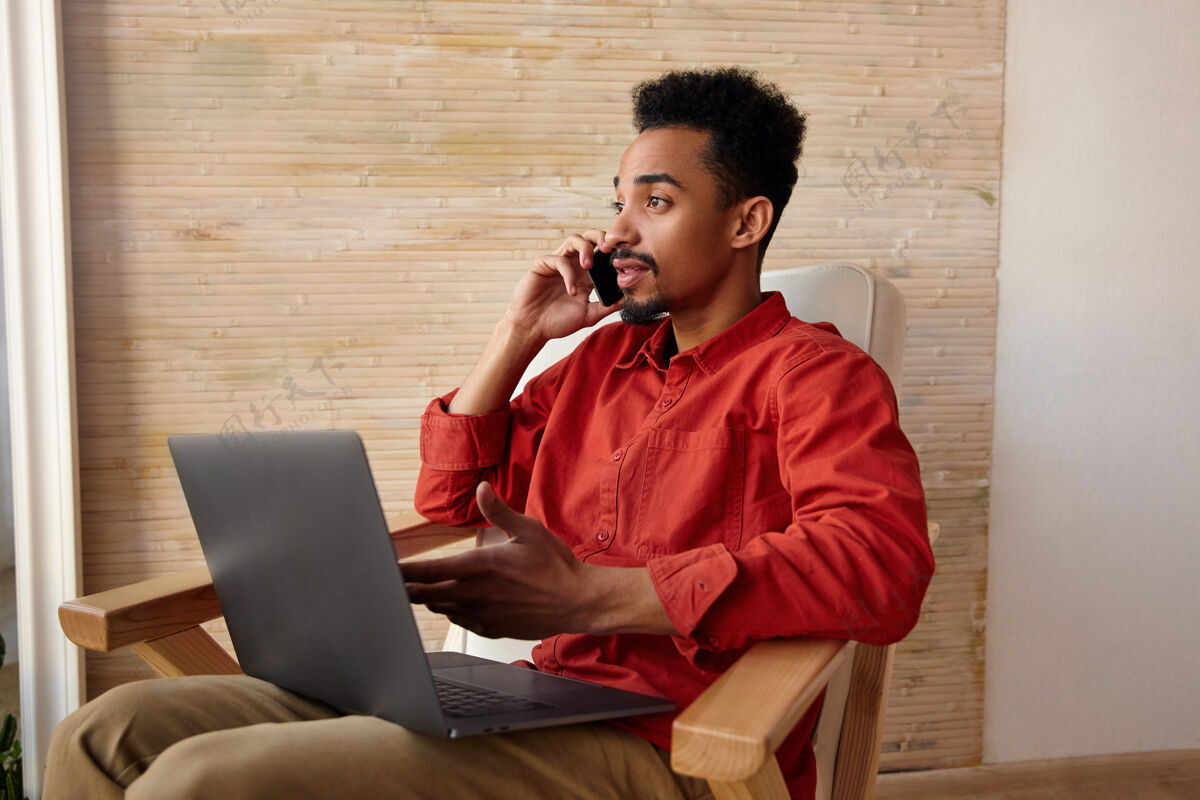 深色一张室内照片 一个年轻的短发留胡子的黑皮肤男人坐在椅子上对着窗户打电话 在办公室外工作摆姿势智能手机表情