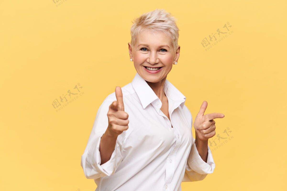 表情快乐迷人的五十岁女性穿着时尚的白衬衫指着前指微笑 选择你与她共舞 面带灿烂的笑容肢体语言华丽微笑展示