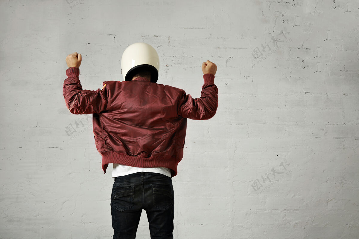 男身穿白色摩托车头盔和勃艮第飞行员夹克的男子在白色墙壁背景下用沙卡手势从背后向上挥舞双拳射击牛仔裤肖像头