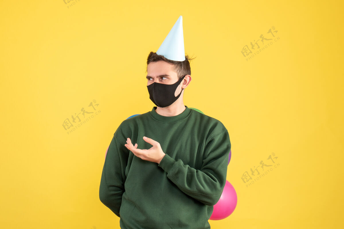 罪犯正面图：戴着派对帽 站在黄色地面上的彩色气球的年轻人肖像成人年轻人