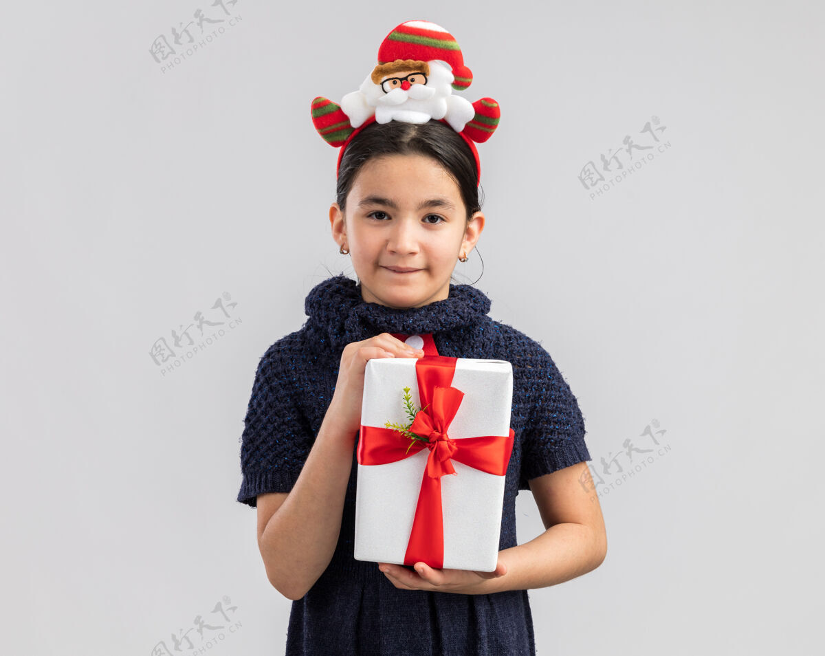 头快乐快乐的小女孩 穿着针织连衣裙 头上系着一条红色领带 头上戴着一个有趣的圣诞圈 手里拿着圣诞礼物 脸上挂着微笑编织礼物圈