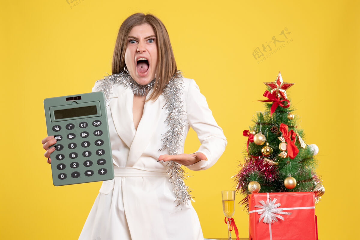新正面图女医生站着 愤怒地拿着黄色背景上的计算器 还有圣诞树和礼品盒站着肖像护士