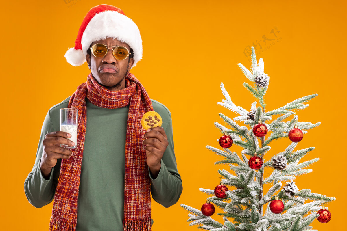 悲伤一个戴着圣诞帽 围着围巾 捧着牛奶和饼干 撅着嘴的悲伤的非洲裔美国人站在橙色背景下的圣诞树旁抱着围巾站着