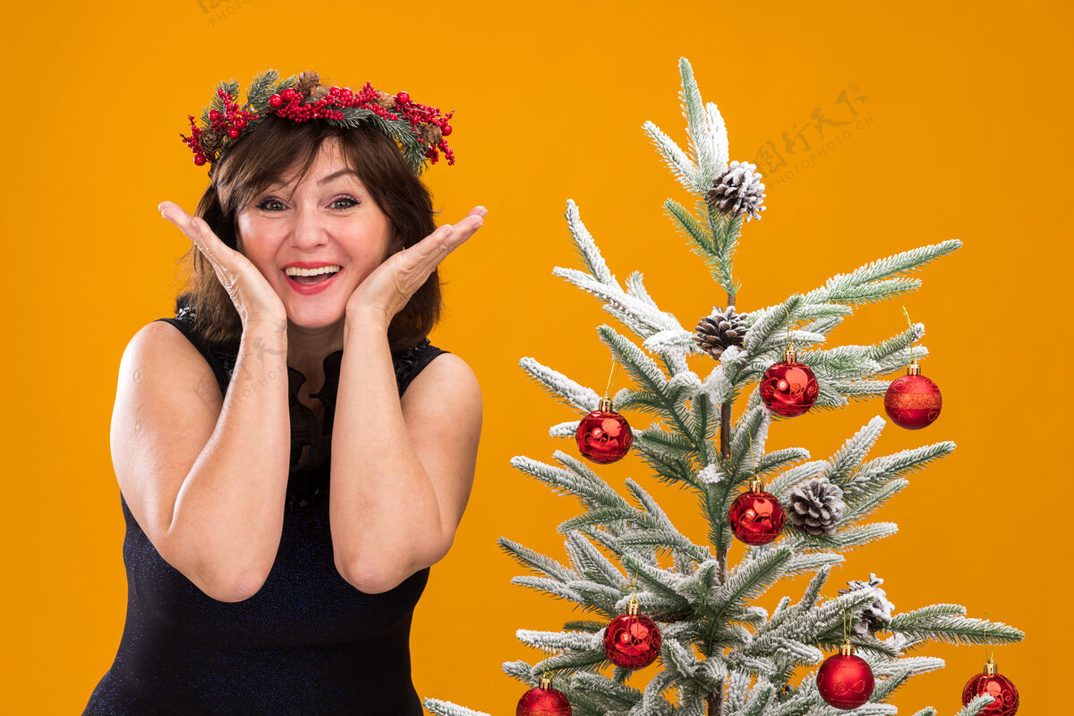 手兴奋的中年妇女头戴圣诞花环 脖子上戴着金属丝花环 站在装饰好的圣诞树旁装饰看附近