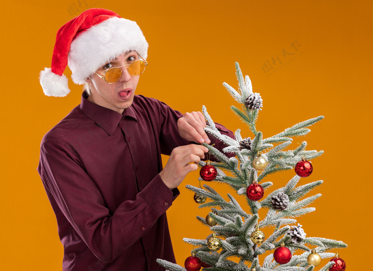 圣诞饰品可爱的金发女郎戴着圣诞帽和眼镜站在橙色背景装饰的圣诞树旁的侧视图中金发站着展示