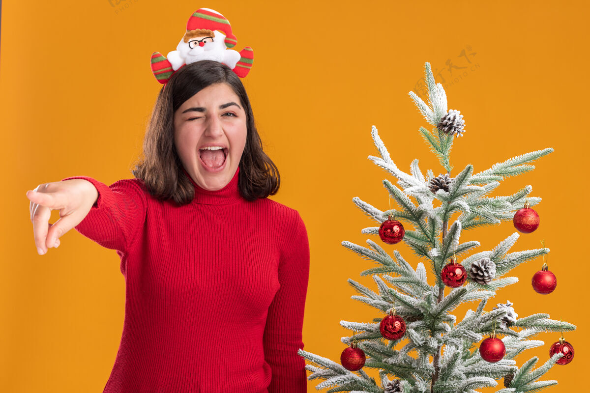 年轻穿着圣诞毛衣戴着滑稽头带的年轻女孩在橙色背景下的圣诞树旁尖叫站着手指指着