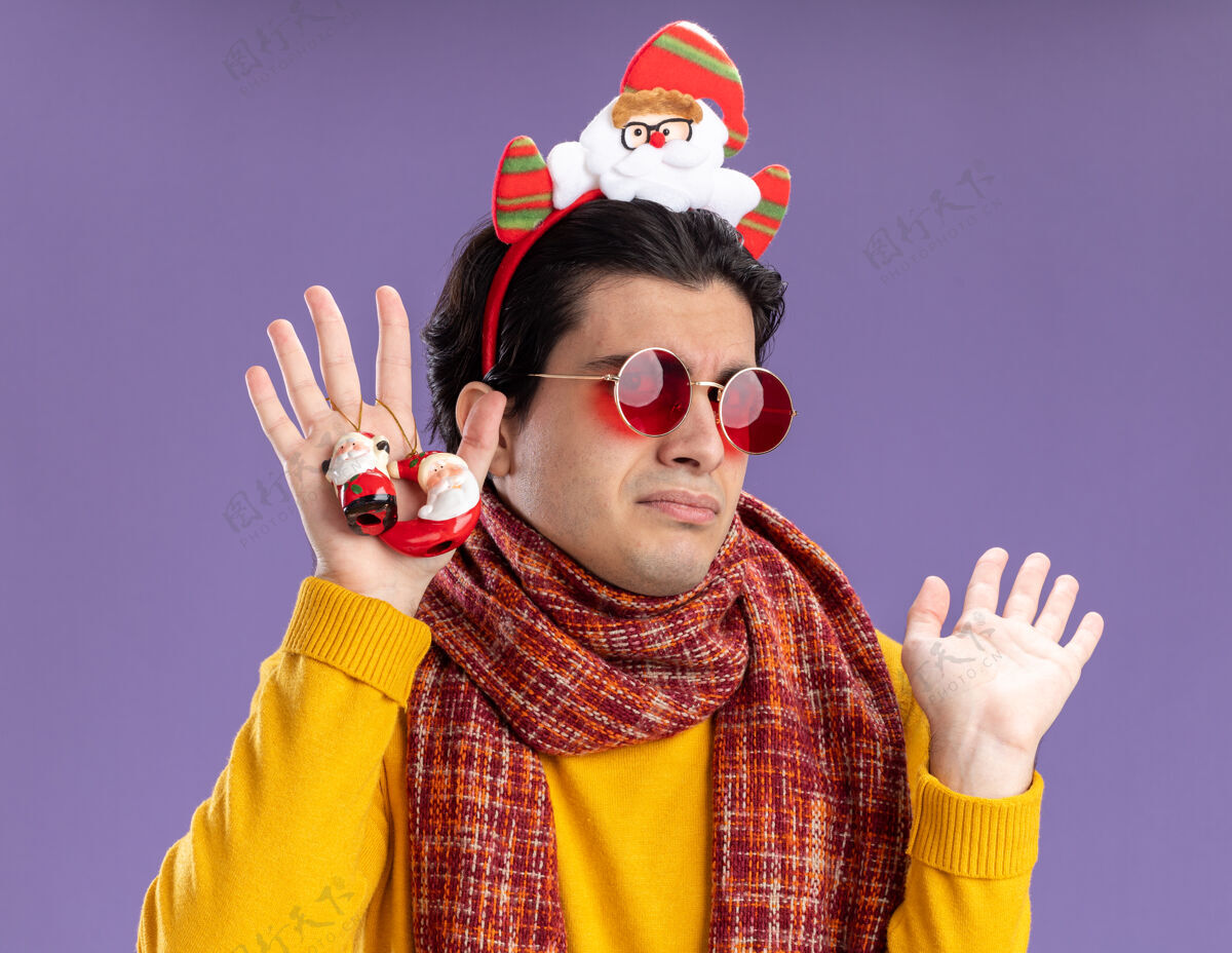 站立脖子上围着暖和的围巾 穿着黄色高领毛衣 头上戴着一副滑稽的眼镜 手里拿着圣诞玩具 站在紫色的墙上 满脸疑惑圣诞节高领毛衣围巾