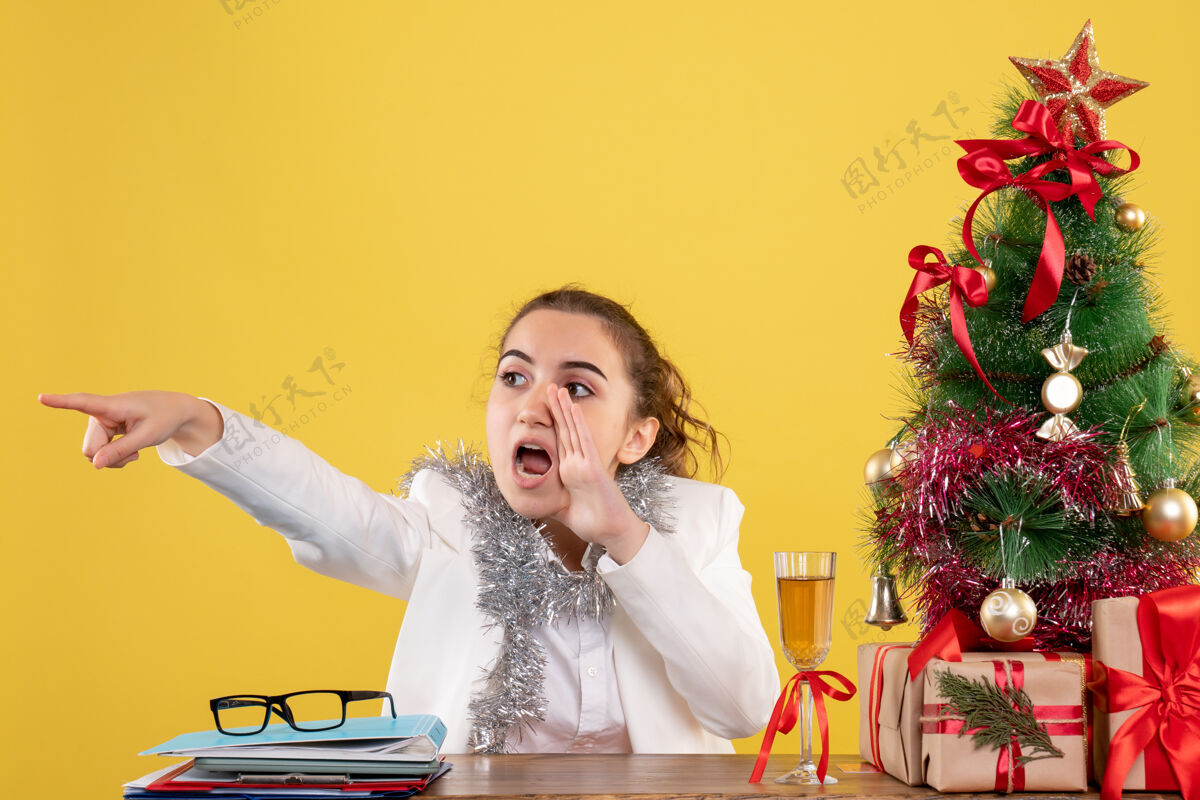圣诞节正面图：女医生坐在黄色背景的桌子后面 还有圣诞树和礼品盒发言人快乐背景