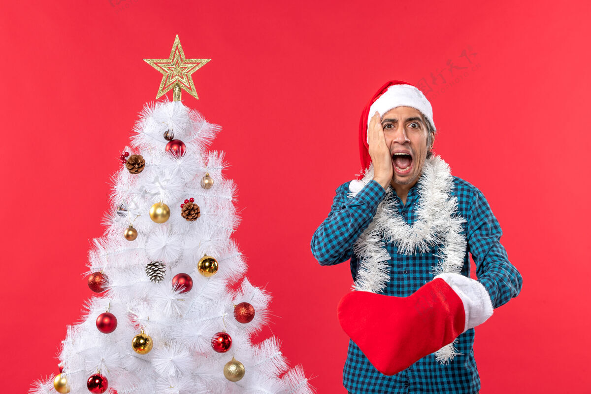 袜子一个戴着圣诞老人帽子 穿着蓝色条纹衬衫 穿着圣诞袜子的震惊的年轻人帽子圣诞圣诞树